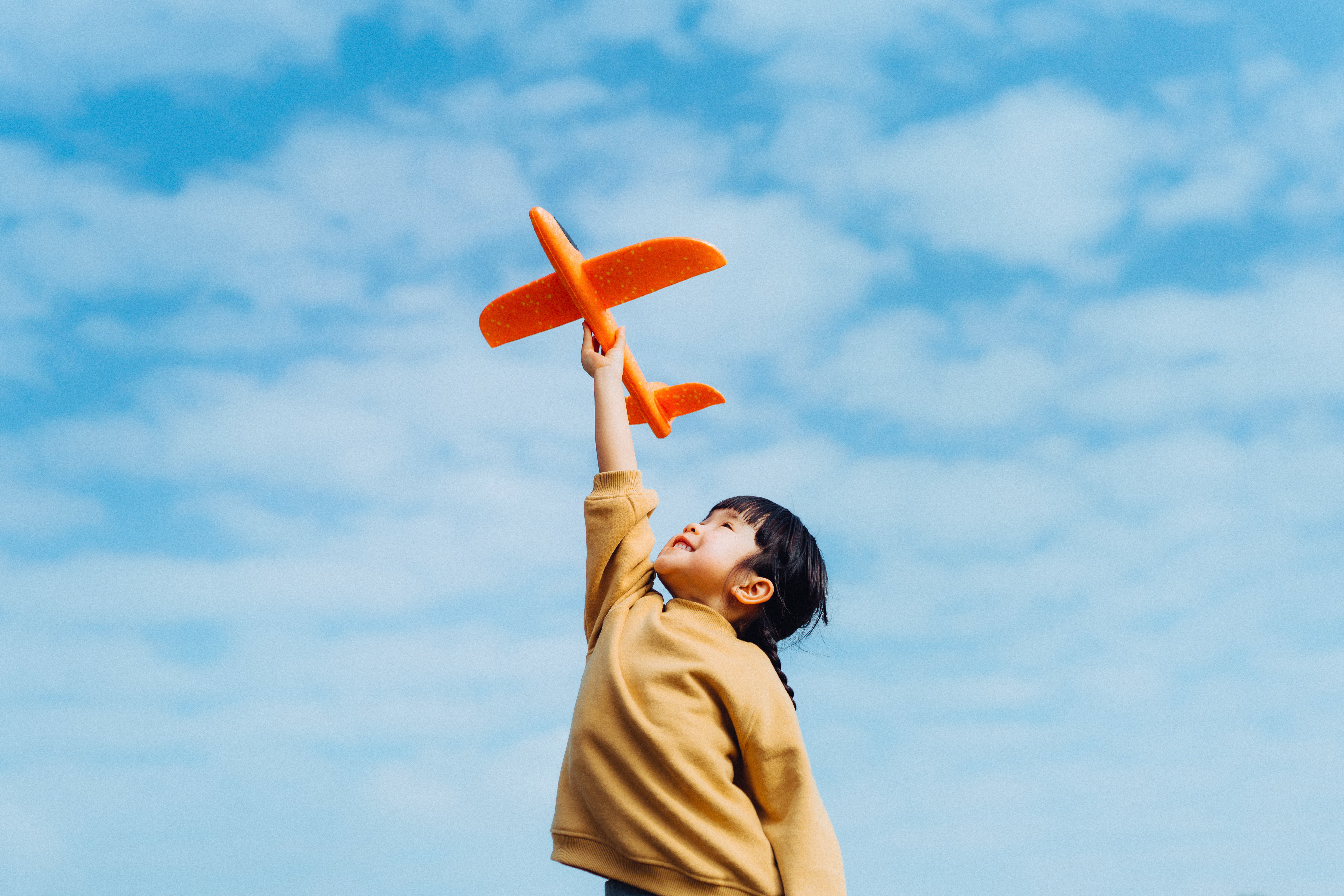 A child holds a red airplane toy above their head in front of a cloudy sky backdrop.