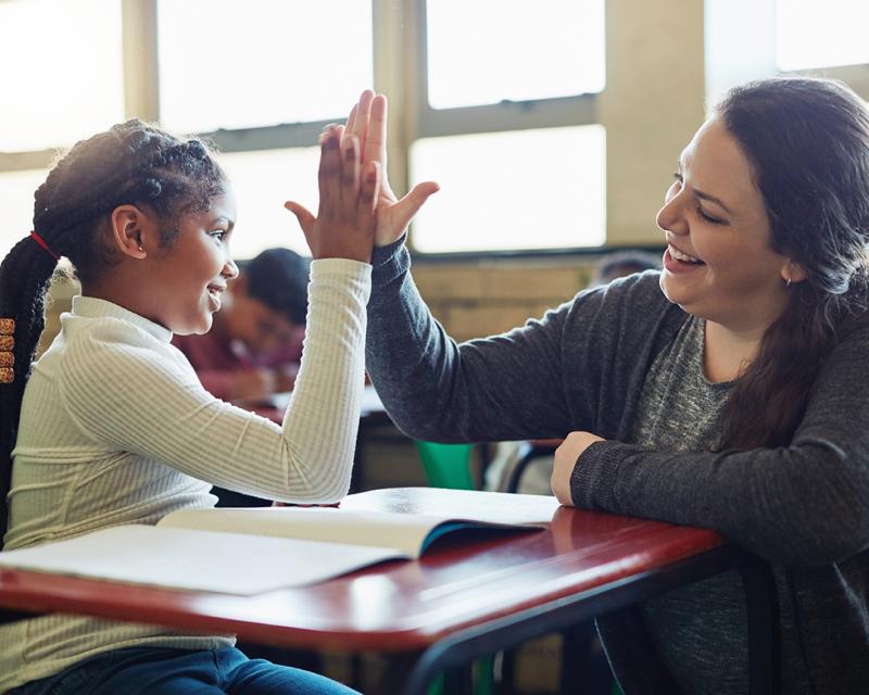 A teacher high-fives a student at a desk. 