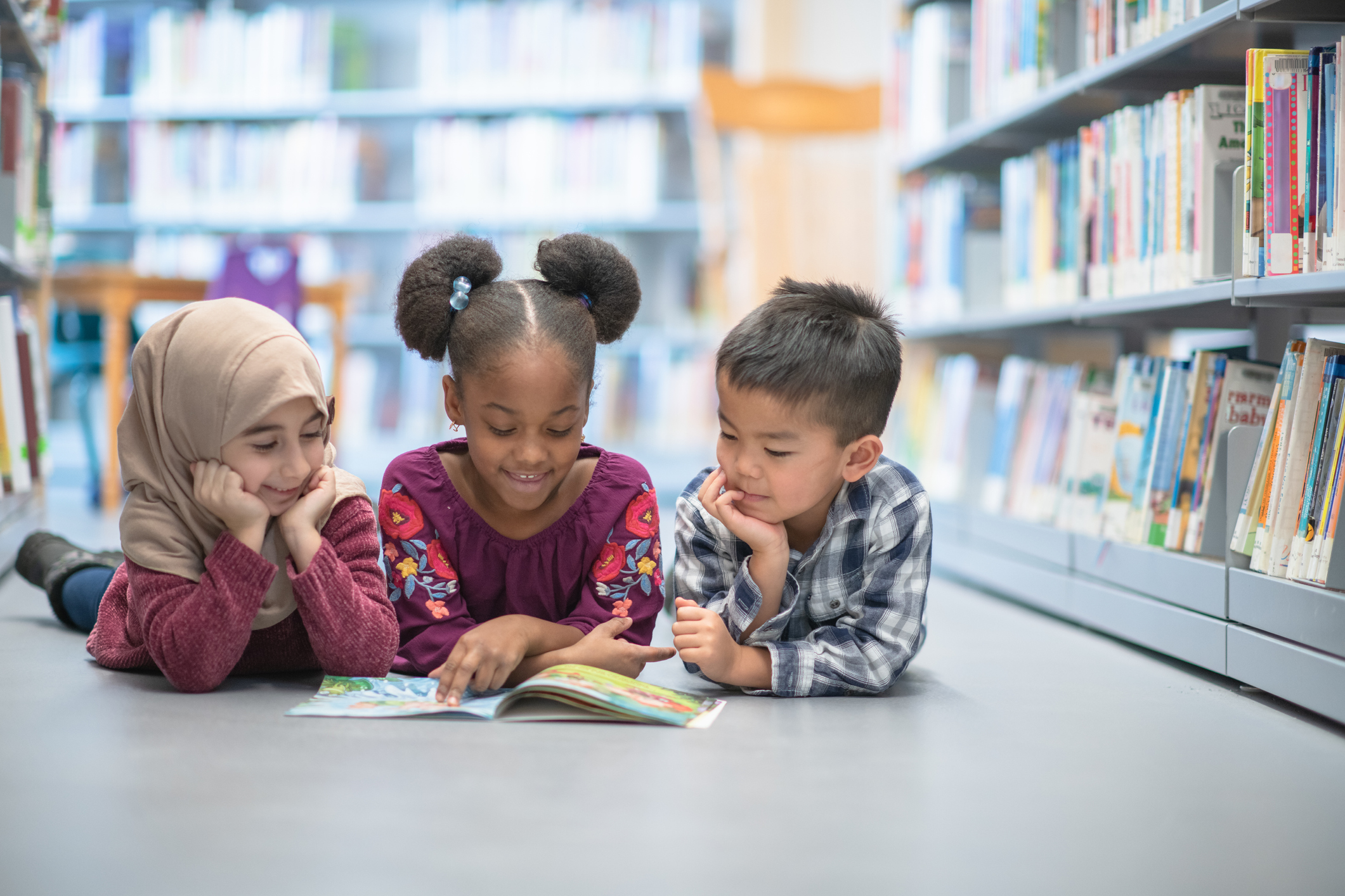 Three children laying on their stomachs between bookshelves, smiling and staring at a shared open book. 