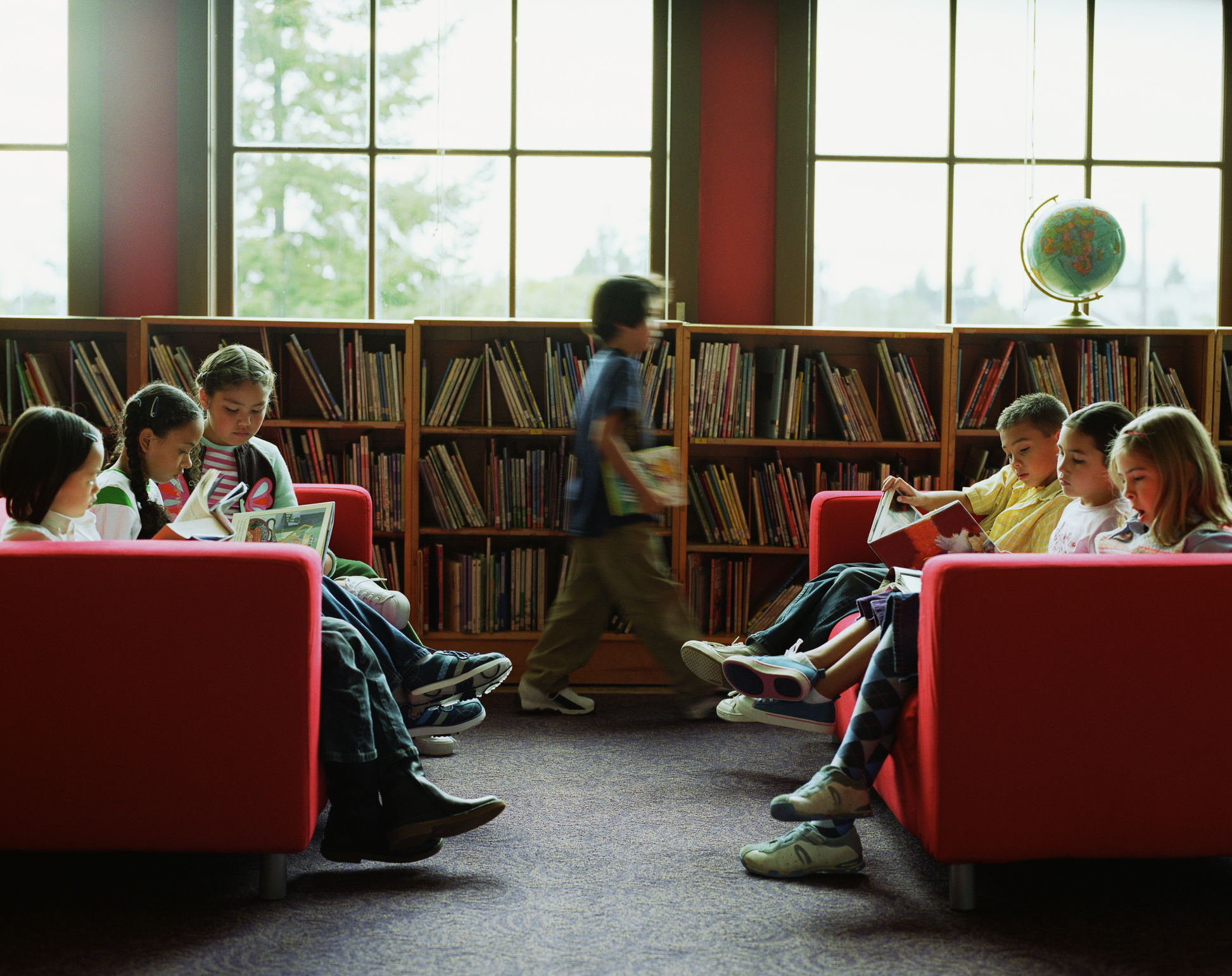 Students in a library sit on couches that face each other reading. A student in the background walks past bookshelves. 