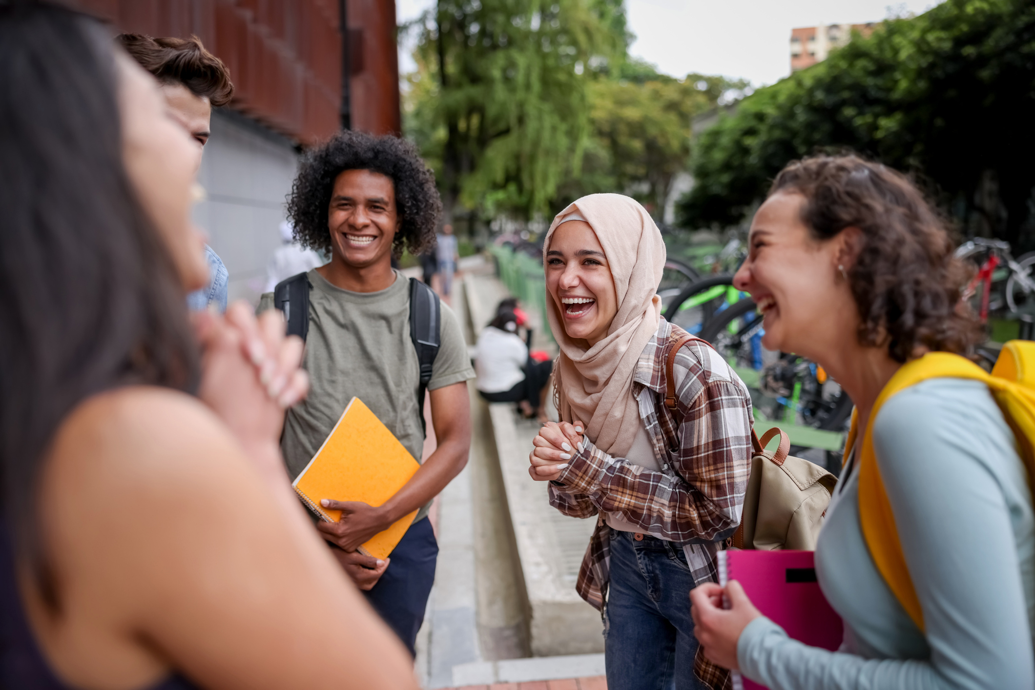 A group of college students stand outside on a college campus wearing backpacks and laughing together. 